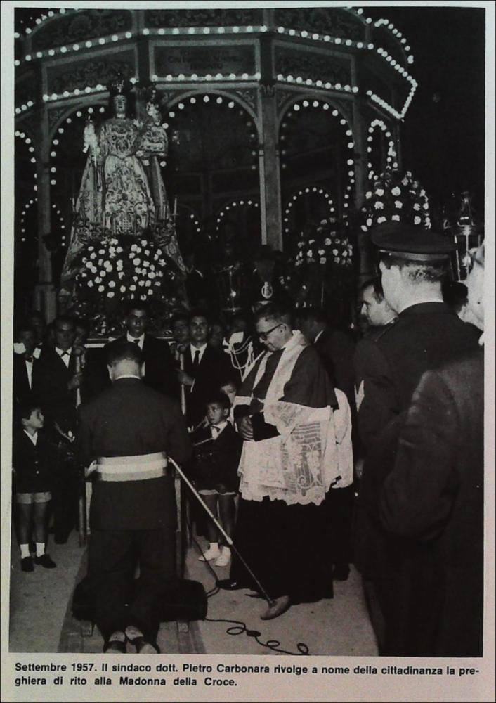 La fotografia storica, risalente al settembre 1957, immortala il momento solenne in cui il sindaco di Triggiano, dott. Pietro Carbonara, eleva alla Madonna la preghiera rituale a nome dell’intera cittadinanza.
La grande statua devozionale della Patrona si staglia maestosa, avvolta da fasci di rose e dallo splendore delle luminarie predisposte in Suo onore, suggellando un momento di intensa partecipazione e profonda devozione collettiva.
Le origini della devozione alla Madonna della Croce risalgono al XVI secolo, quando, fuori dall’antico abitato, venne edificata un’edicola sacra decorata con un’immagine “miracolosa” ad opera dei cosiddetti “homini de fora”, ossia contadini e braccianti che ogni giorno passavano di lì per andare a lavorare nei campi.
Una guarigione riten...
