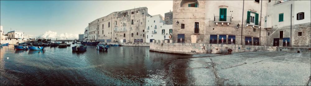 Lo scatto riesce a catturare in un solo sguardo tutto il porticciolo vecchio di Monopoli con il suo lungo passaggio sotto le mura degli antichi palazzi che raggiunge il molo. Le mura sono allestite con gli scatti di Mustafa Sabbagh (Amman, Giordania, 1961) del progetto “Corpus fugit”. I soggetti sono giovani ragazzi e ragazze che immortalati su uno sfondo scuro mettono in luce la loro individualità....