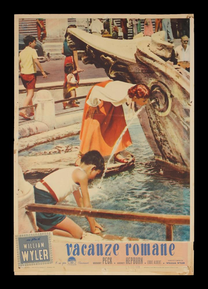 L'immagine riproduce uno dei manifesti conservati presso la Mediateca Regionale Pugliese (vedi Note). Si tratta di una delle fotobuste del film “Vacanze romane”, del regista William Wyler, girato interamente a Roma. Si tratta di un soggetto su supporto cartonato dalle misure 35x50 che immortala uno scatto di scena del film: la bellissima Hepburn mentre beve in una fontana di Roma. Nella parte inferiore abbiamo il titolo in corsivo minuscolo, il nome degli attori principali, del regista ed i credits....