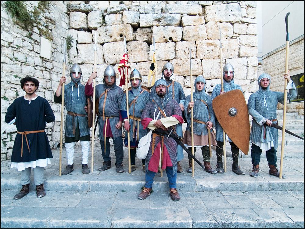 L'immagine digitale è parte di una raccolta fotografica che documenta la seconda edizione della Festa di San Rocco in Contea svoltasi nel 2007 a Conversano e curata dall'associazione culturale “I Collegia”. L'autore della fotografia ritrae un gruppo di cavalieri normanni ovvero figuranti facente parte dell'associazione “Compagnia d'Arme Stratros”. Con la spada in una mano e lo scudo nell'altra, i figuranti indossano gli elementi iconografici distintivi dei soldati di quel tempo come le scarpe in pelle senza lacci e la suola in cuoio, l'elmo e il camaglio in maglia di ferro per proteggere il capo. Durante la rievocazione storica di San Rocco l'associazione Stratos ha curato la ricostruzione di un accampamento militare nonché la simulazione di alcune scene di vita militar...
