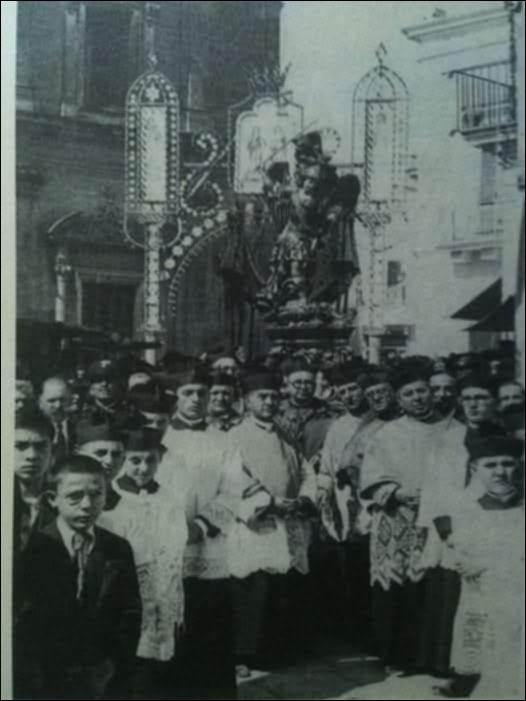 Questa foto storica, datata al 1945, immortala la monumentale statua del Patrono di Massafra San Michele Arcangelo in partenza per la processione per le vie del paese in occasione della Festa Patronale.
Il culto di San Michele, ufficializzato nel 1721 ma presente già da secoli, affonda le sue origini in antiche devozioni legate anche alla storica grotta carsica dedicata all’Arcangelo in zona Varcaturo, luogo di culto e testimonianza di riti e credenze tra epoche preclassiche e cristiane. Raffigurato come guerriero celeste, simbolo di protezione e giustizia, il santo è al centro di riti, opere d’arte, leggende e pratiche popolari che si tramandano di generazione in generazione, rafforzando il senso di appartenenza e coesione sociale.
Nonostante periodi di sospensione, la...