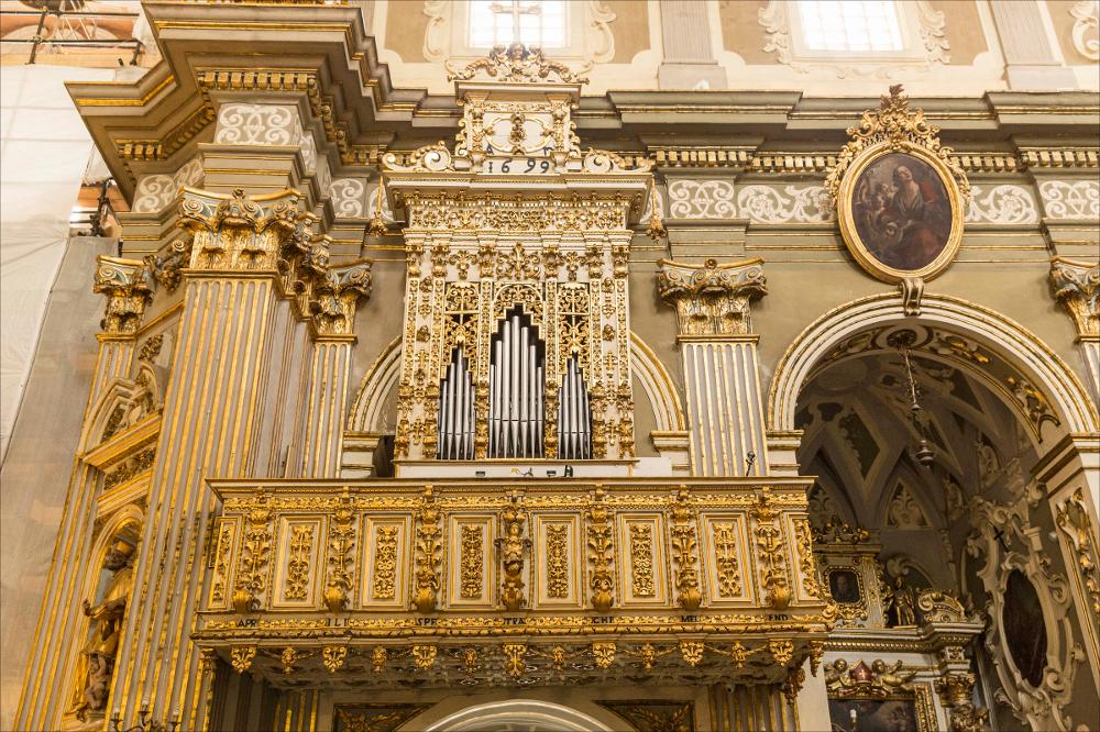 L’organo, qui raffigurato nell'immagine digitale, è collocato nella chiesa del Crocifisso di Galatone, in provincia di Lecce. L’edificio, costruito intorno al 1683, è a croce latina con tre cappelle per ogni lato della navata, fu realizzato grazie all’apporto di fra Nicolò da Lequile (Leonardo Milelli) e dell’architetto leccese Giuseppe Zimbalo e consacrato nel 1711 dal vescovo Antonio Sanfelice. La monumentale facciata è divisa in tre ordini. Nel primo ordine un fascio di lesene, sormontate da capitelli corinzi, scandiscono il paramento murario con quattro Santi collocati in eleganti nicchie decorate. Il portale centrale invece presenta al centro della trabeazione una scultura a mezzo busto di Cristo Crocifisso, sormontato da un curioso baldacchino sostenuto da putti. ...