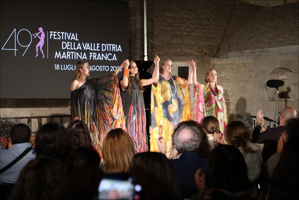 <div>Durante la serata del ciclo <em>Il Canto degli Ulivi</em> del 27 luglio 2023, presso la <strong>Masseria Nicola Casavola</strong> a Martina Franca, quattro cantanti dell’<strong>Accademia del Belcanto “Rodolfo Celletti”</strong> salutano al termine della loro esibizione. L’evento, inserito nella 49ª edizione del <strong>Festival della Valle d’Itria</strong>, ha valorizzato la tradizione musicale della regione, coniugando l’eccellenza artistica alla suggestiva cornice storica della masseria, un edificio settecentesco immerso in oliveti secolari, e al paesaggio unico della Valle d’Itria.</div>...