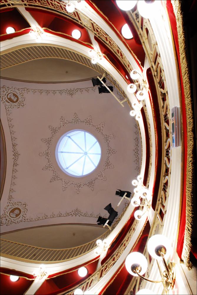 L'immagine è parte della campagna fotografica realizzata dalla fotografa Margherita Mirabella per Teatro Pubblico Pugliese, che ha ripreso nei suoi scatti le eleganti architetture e i rifiniti dettagli di alcuni dei più bei teatri pugliesi e alcuni degli spettacoli teatrali portati in scena in questi ultimi anni. Scattata dal basso, la fotografia mostra parte dei palchi, il soffitto decorato e con punto luce del Teatro Comunale di Nardò, situato in Via Mauro Manieri, piccolo gioiello culturale ottocentesco della città salentina. Dopo l'Unità d'Italia, l'esigenza di avere un Teatro Comunale per la popolazione locale spinse la Prefettura ad affidare l'incarico per la sua realizzazione. Nel 1893, fu accolto il progetto dell'Ing. Quintino Tarantino e i lavori iniziarono nel...