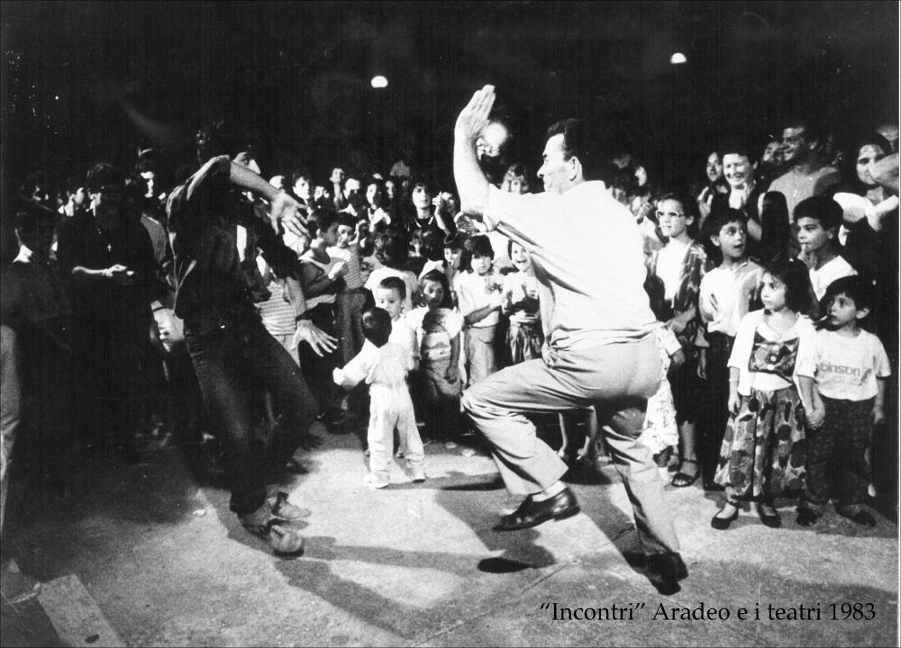L'immagine digitale immortala un piccolo momento della rassegna “Aradeo e i Teatri” inaugurata nello stesso anno, 1983, con il titolo “Incontri”, un appuntamento con il teatro sperimentale. La manifestazione fu organizzata dal circolo culturale “S.M. Ejzenstein” per colmare, attraverso la messa in scena di spettacoli di teatro e danza, la mancanza di confronto fra culture differenti molto sentita dalla cittadinanza di Aradeo. Nello specifico si osserva un particolare momento della festa organizzata nella piazzetta Municipio dove si alternano danzatori di pizzica e di scherma. La "pizzica a scherma" è spesso impropriamente chiamata "danza delle spade” nonostante l'arma utilizzata sia il coltello, rappresentato figurativamente dal dito indice e medio della mano. La perfor...
