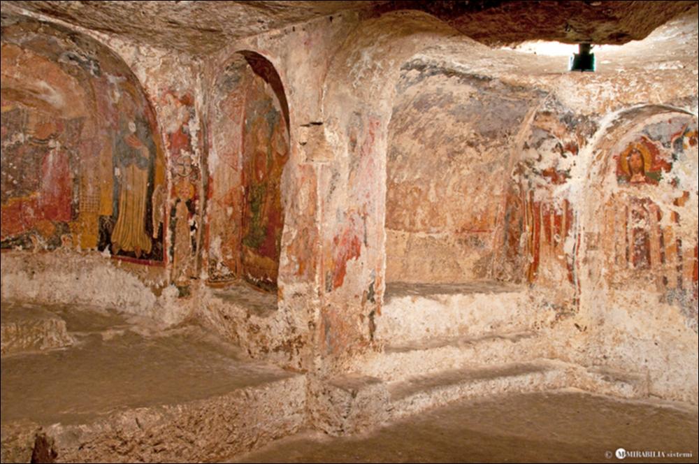 L'immagine digitale rappresenta un particolare dell'interno della chiesa rupestre dedicata a Sant'Antonio Abate, sita sotto l'ex ospedale Pagliari di Massafra. Il complesso, nel corso del tempo, ha assunto diverse funzioni: legnaia, deposito, vasca per la calce viva ed altro. Verso la fine degli anni Cinquanta del secolo scorso, volontari della Pro Loco massafrese si sono impegnati nello sgombero dell'area, riportando alla luce gli affreschi residui, alcuni dei quali sono stati restaurati dalla Soprintendenza. Ricavata dall'accorpamento di due chiese distinte, officiate – probabilmente – una secondo il rito orientale ed una secondo il rito latino, è un esempio classico di chiesa ipogeica. Tra le pareti è possibile notare numerosi affreschi risalenti a diversi periodi, d...