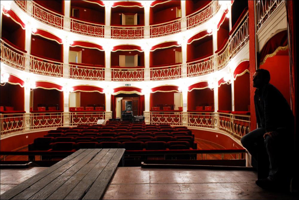 L'immagine è parte della campagna fotografica realizzata dalla fotografa Margherita Mirabella per Teatro Pubblico Pugliese, che ha ripreso nei suoi scatti le eleganti architetture e i rifiniti dettagli di alcuni dei più bei teatri pugliesi e alcuni degli spettacoli teatrali portati in scena in questi ultimi anni. Scattata dal palcoscenico, la suggestiva fotografia mostra parte dei palchi e della platea del Teatro Comunale di Nardò, situato in Via Mauro Manieri, piccolo gioiello culturale ottocentesco della città salentina. Dopo l'Unità d'Italia, l'esigenza di avere un Teatro Comunale per la popolazione locale spinse la Prefettura ad affidare l'incarico per la sua realizzazione. Nel 1893, fu accolto il progetto dell'Ing. Quintino Tarantino e i lavori iniziarono nello ste...
