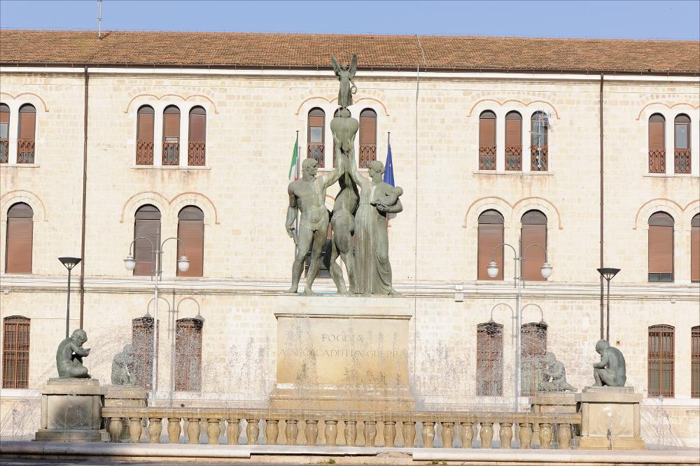 L'immagine digitale mostra il Monumento ai Caduti a Foggia, opera dello scultore napoletano Amleto Cataldi formatosi tra la bottega del padre Angelo, intagliatore del legno, e la scuola romana di Ripetta. La scultura risale al 1928 e fu posizionata al centro della odierna Piazza Italia. Si tratta di una tipologia di monumento unica in Puglia, una fontana quadrangolare con un recinto a balaustrata e agli angoli quattro putti in bronzo accovacciati ed intenti a soffiare in una conchiglia. Al centro, un basamento in pietra accoglie tre figure in bronzo: due eroi e una donna che sostiene un neonato, tutti reggono un cuore su cui è collocata una figuretta alata. I due eroi raffigurati con una possente muscolatura mostrano fieri il loro corpo: uno presenta un'armatura elegant...