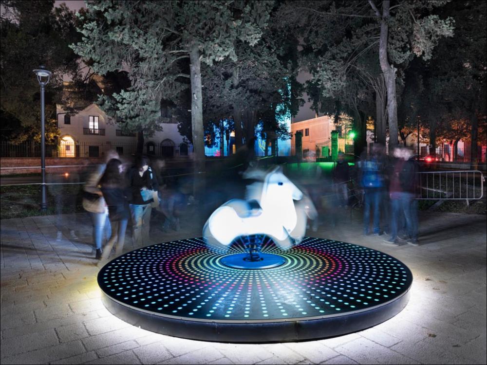 Lo scatto fotografico riprende “Never Ends”, un’installazione interattiva degli artisti italiani Luigi Console e Valentina Novembre, collocata presso Villa Donnaloja ad Alberobello. Al centro della scena un cavalluccio luminoso su molla, emblema del gioco, dell’allegria e della fantasia. Il cavalluccio era pronto a illuminarsi nel momento in cui il visitatore ci saliva su, accendendo luci sulla pedana sottostante e creando un’ambientazione fiabesca, stimolo a riscoprire il piacere del gioco e del “sentirsi bambini”.
L’installazione è stata realizzata nell’ambito della seconda edizione del Life - Alberobello Light Festival 2017 che ha illuminato il centro cittadino e alcuni dei suoi luoghi più caratteristici dal 30 settembre all’8 ottobre.
Il Festival è organizzato da Li...