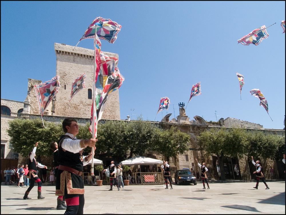 L'immagine digitale è parte di una raccolta fotografica che documenta la seconda edizione della rievocazione storica di San Rocco svoltasi nel 2007 a Conversano e curata dall'Associazione culturale “I Collegia”. Dal 2006, la tradizionale Cavalcata si è arricchita del corteo che unisce alla tradizione devozionale la bellezza di elementi laici come fuochi pirotecnici, bande, luminarie, i giochi degli artisti di strada e la compagnia di sbandieratori e musici. La parata parte da Piazza Castello, attraversa il centro storico di Conversano per toccare i principali punti della città, rievocando pratiche devozionali del passato e anche storie e leggende legate alla Badessa Maria d'Angiò. Scattata in una situazione di luce piena, da una posizione un po' bassa, la fotografia dig...