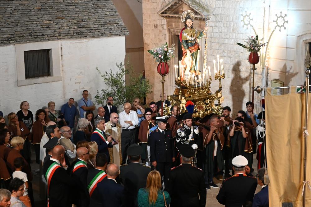 La processione di San Rocco a Noci attraversa le vie del paese, con la statua del Santo sorretta dai volontari vestiti con la caratteristica mantella marrone, simbolo di devozione e servizio. Dietro di loro si scorge l’edificio delle Monache di Santa Chiara, conosciuto a Noci come il Chiostro delle Clarisse o l’ex sede delle Monache di Santa Chiara. Si tratta di un edificio storico di origine seicentesca, parte dell’antico monastero delle monache clarisse, che è stato ristrutturato e oggi costituisce un luogo di interesse nel centro del paese.<div><br>
La festa patronale si celebra tradizionalmente il 16 agosto, giorno dedicato al Santo, mentre la solennità liturgica propria della comunità nocese ricorre nella prima domenica di settembre, cuore delle celebrazioni religi...