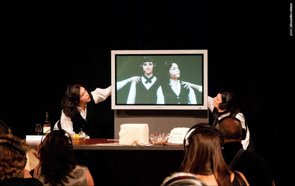 L'immagine in oggetto, fotografia digitale ad opera di Alessandro Colazzo, cattura un momento di “Il pasto della Tarantola”, spettacolo dei Cantieri Teatrali Koreja di Lecce, che fa parte della loro produzione originale e annuale. Con un format originalissimo e all'avanguardia, la rappresentazione teatrale interpretata, da Ottavia Perrone e Maria Rosaria Ponzetta, punta a coinvolgere lo spettatore nella scoperta dei sapori tipici salentini. Mùscari, scapèce, pomodori secchi sott'olio, africani e il vino negramaro ornano una tavola dove una delle attrici è distesa silenziosamente, mentre l'altra comincia il racconto dei prodotti tipici e guida il pubblico in una degustazione individuale. Il Salento emerge in tutta la sua forza e fascino, anche nel racconto di un fenomeno...