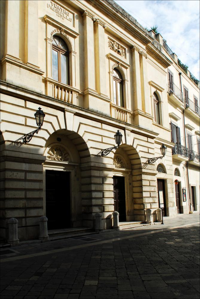 L'immagine è parte della campagna fotografica realizzata dalla fotografa Margherita Mirabella per Teatro Pubblico Pugliese, che con i suoi scatti ha ripreso le eleganti architetture e i rifiniti dettagli di alcuni dei più bei teatri pugliesi e di alcuni spettacoli teatrali portati in scena in questi ultimi anni. La fotografia, scattata all'esterno del Teatro Paisiello di Lecce, ci mostra come appare esternamente la struttura teatrale progettata dall'architetto Oronzo Bernardini e sorta su una preesistente costruzione in legno risalente al XVIII secolo. Lineare e sobria, la facciata in pietra locale custodisce i simboli del teatro classico greco. Nel 1870 il teatro, dedicato al musicista tarantino Giovanni Paisiello, viene inaugurato con la rappresentazione dell'opera “I...