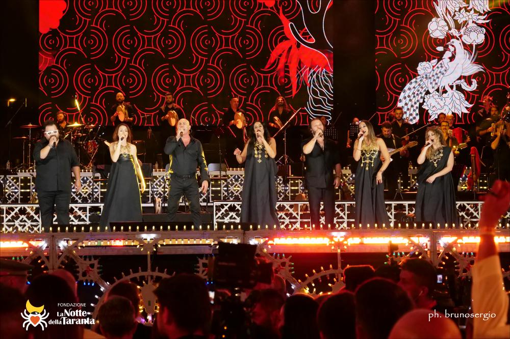 <div>Lo scatto mostra l’orchestra della Notte della Taranta con il gruppo voci al completo al centro del palco, mentre sullo sfondo si staglia la scenografia digitale. Composto da cantanti professionisti provenienti dal Salento, il gruppo interpreta i brani della tradizione popolare locale. L’immagine documenta l’armonia tra strumenti, voci e tecnologia scenica che caratterizza l’edizione 2022 del festival.</div>...