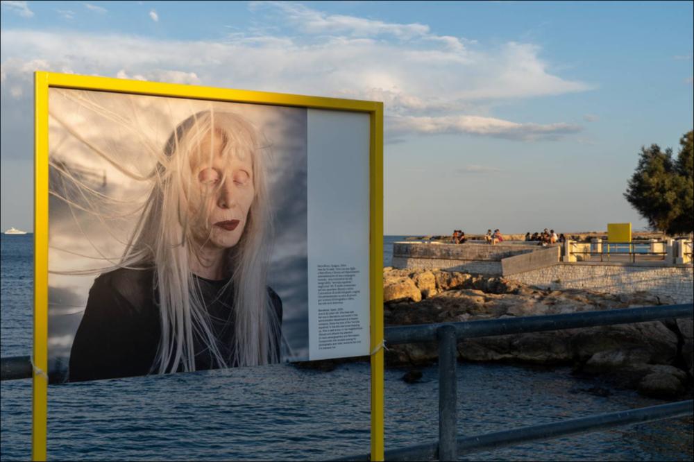 Presso il Belvedere di Porta Vecchia, le luci del tramonto incorniciano i pannelli fotografici del progetto “Albino Beauty” di Paola De Grenet, dove la pelle perlacea diventa una tela per ricchi punti di colore dati dai vestiti e dal trucco delle sue modelle. Paola De Grenet è una fotografa italiana (nata a Milano, 1971) di base a Barcellona, specializzata in ritrattistica e reportage. Dopo gli studi di grafica a Londra, inizia la sua carriera come fotografa nel 1999. Il suo progetto vuole mettere in contatto diretto l'osservatore con cosa fa gli paura e non conosce, con lo scopo di comprenderlo e rispettarlo. I ritratti degli albini celebrano una tipologia di bellezza unica, eliminando il limite della differenza. 
“Il loro aspetto pallido e delicato e la loro condizion...