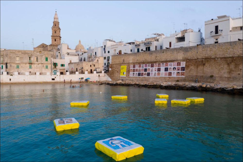 Nella marina di cala Porta vecchia, la mostra fotografica di Tadao Cern (Lituania, 1983) diventa una vera installazione artistica che non solo coinvolge l’osservatore, ma anche interagisce con lo spazio marino sotto forma di galleggianti dove sono stampate le sue opere, fotografie dall’alto di bagnanti. 
Secondo Tadao Cern, la spiaggia è l’unico luogo dove gli esseri umani si lasciano analizzare spontaneamente, diventando l’occasione per approfondirne le differenze e studiarne le abitudini naturali. La scelta dei galleggianti permette di moltiplicare l’azione fotografata dall’artista essendo possibile salirci sopra e sdraiarsi....
