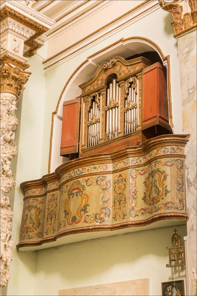 L’organo raffigurato nell'immagine digitale in oggetto è collocato nella chiesa di S. Leonardo a Manduria. La chiesa di piccole dimensione fu costruita su una preesistente cappella intitolata alla Madonna di Costantinopoli. Dedicata a S. Leonardo dal 1621, venne ricostruita a partire dal 1702, grazie ai contribuiti dei confratelli. La facciata molto semplice presenta doppie paraste ai lati e un fastigio a volute, collocato su una trabeazione continua, sormontato da una croce. Il portale centrale, unico ingresso, vede al centro all’interno una nicchia con la statua di San Leonardo, ai lati San Cristoforo e San Raffaele. L’interno ad aula unica presenta sull’altare maggiore, realizzato in pietra leccese e con colonne tortili, la tela della Madonna di Costantinopoli realiz...