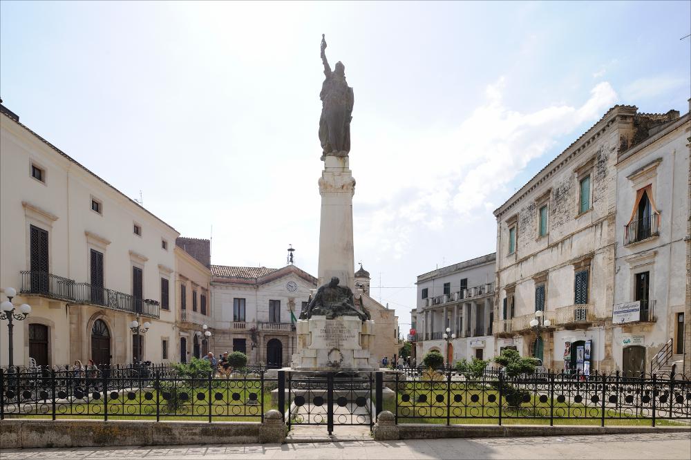 L'immagine digitale mostra l'insieme del Monumento ai Caduti di Grumo Appula. Posto al centro di Piazza Vittorio Veneto, il monumento è in pietra, marmo e bronzo di tipo "a cippo" e fu realizzato dallo scultore barese Mario Sabatelli negli anni '20, su commissione dell'Amministrazione comunale cofinanziato dalla comunità dei conterranei residenti negli Stati Uniti con 35.000 Lire. Fu scelta, come si evince dalle fonti dell'epoca, l'opera del Sabatelli perché rispecchiava i valori storici e filosofici richiesti dalla committenza e il profondo senso di riconoscenza verso i Caduti e il trionfo della Vittoria. In foto, si riconoscono i due gruppi scultorei in bronzo che ornano il monumento: in basso la raffigurazione dei Caduti, mentre in alto spicca l'Allegoria della Patri...