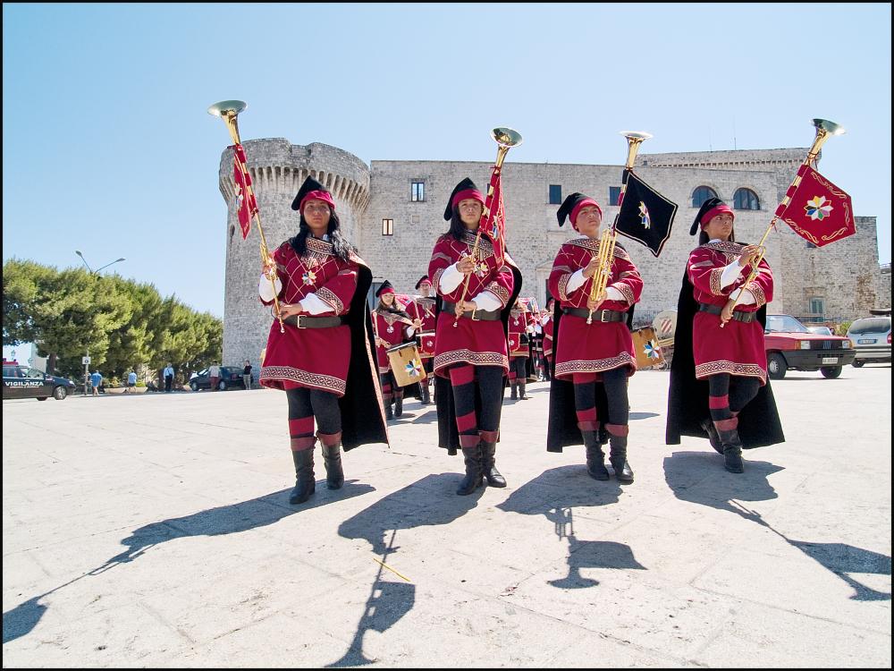 L'immagine digitale è parte di una raccolta fotografica che documenta la seconda edizione della rievocazione storica di San Rocco svoltasi nel 2007 a Conversano e curata dall'Associazione culturale “I Collegia”. Dal 2006, la tradizionale Cavalcata si è arricchita del corteo che unisce alla tradizione devozionale la bellezza di elementi laici come fuochi pirotecnici, bande, luminarie, i giochi degli artisti di strada e la compagnia di sbandieratori e musici. Il corteo parte da Piazza Castello, attraversa il centro storico di Conversano per toccare i principali punti della città, rievocando pratiche devozionali del passato e anche storie e leggende legate alla Badessa Maria d'Angiò. Scattata in una situazione di luce piena, da una posizione un po' bassa, la fotografia dig...