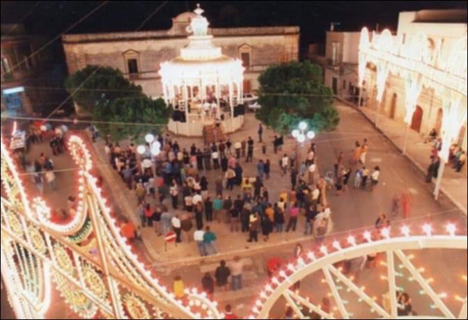 <div class="ql-editor"><div><strong>Foto storica del 1970. </strong>Scatto dall’alto che ritrae Piazza Municipio a Villa Castelli durante la Festa Patronale. Al centro domina la cassarmonica splendidamente illuminata, rendendo la piazza un vero teatro a cielo aperto. La folla si avvicina alla struttura con passo lento e curioso, stringendosi attorno per assistere all’esibizione musicale. Dall’alto si percepisce il movimento compatto delle persone, l’attesa condivisa, il richiamo irresistibile della musica che raduna generazioni diverse sotto lo stesso cielo di festa.</div><div><br></div><div>La Festa Patronale di <strong>Villa Castelli</strong> si svolge ogni anno dall’1 al 3 ottobre ed è uno degli appuntamenti più attesi e partec...