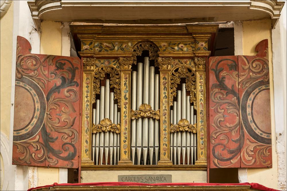 L’organo raffigurato nell'immagine digitale è collocato nella chiesa del Carmine di Manduria. La chiesa viene fondata dagli Scolopi nel 1681 da Giacomo Antonio Carrozzo. I lavori di costruzione iniziarono nel 1701 e terminarono nel 1741 (come ricorda la targa nel timpano della facciata), mentre il convento fu terminato nel 1745. La facciata a due ordini, presenta nel piano inferiore un portale monumentale con un timpano triangolare spezzato, ai lati i due ingressi minori sormontati da due nicchie con le statue delle sante Irene e Barbara. Il secondo piano diviso da una balaustra, su cui sono collocate le statue degli Evangelisti, è leggermente arretrato, così da costituire uno scenografico affaccio sulla piazza. Inizialmente la chiesa era intitolata ai Santi Pietro e Pa...