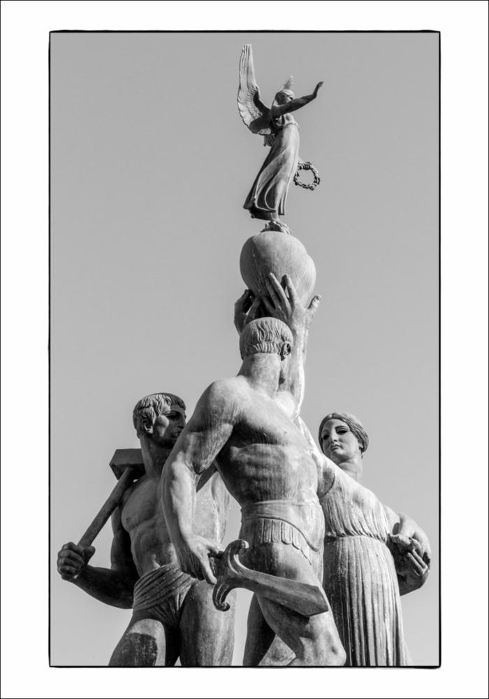 L'immagine digitale mostra un particolare del Monumento ai Caduti a Foggia, opera dello scultore napoletano Amleto Cataldi formatosi tra la bottega del padre Angelo, intagliatore del legno, e la scuola romana di Ripetta. La scultura risale al 1928 e fu posizionata al centro della odierna Piazza Italia. Protagonisti del dettaglio sono tre figure in bronzo: due eroi e una donna che sostiene un neonato, tutti reggono un cuore su cui è collocata una figuretta alata. I due eroi raffigurati con una possente muscolatura mostrano fieri il loro corpo: uno presenta un'armatura elegante, l'altro il corpo completamente nudo, fasciato sola da un perizoma. La figura maschile con l'armatura sostiene una spada e richiama il modello iconografico del militare romano, mentre l'altra figur...