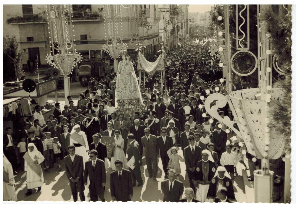 <div>Una foto storica mostra in primo piano la maestosa statua della Madonna del Rosario portata in processione durante la Festa Patronale di Ginosa nell’anno 1957.</div><div>Questa festa, forte di una tradizione plurisecolare di oltre 260 anni, prende avvio nove giorni prima della prima domenica di ottobre con la novena nella Chiesa Matrice di Santa Maria del Rosario.</div><div>Il venerdì precedente la prima domenica di ottobre si svolge la suggestiva processione del quadro della Madonna per le vie del centro cittadino, accompagnata dalla Banda e dalla Bassa Musica di Ginosa. In questa occasione avviene la prima accensione ufficiale delle luminarie artistiche e, in piazza, l’immagine viene collocata sulla cassa armonica per la benedizione dei fedeli, a...