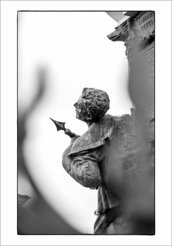 L'immagine digitale mostra un particolare del Monumento ai Caduti ad Ascoli Satriano, opera dello scultore bolognese Giulio Barberi. Racchiuso da una cancellata in ferro battuto, su un doppio gradone, vi è un blocco in marmo sul quale si erge un gruppo bronzeo, sul quale a sua volta si innalza un obelisco su cui sono state inserite delle foglie di palma in bronzo. Il gruppo scultoreo consta di due protagonisti: a sinistra un bersagliere è ritratto nel momento di avanzamento alla carica con moschetto e baionetta innestata, mentre sulla destra, come si può vedere dal dettaglio, vi è un fante colpito che cade avendo tra le braccia la bandiera. I due personaggi sembrano sporgersi da una parete rocciosa. Sui lati dell'obelisco sono presenti delle targhe bronzee: una in facci...