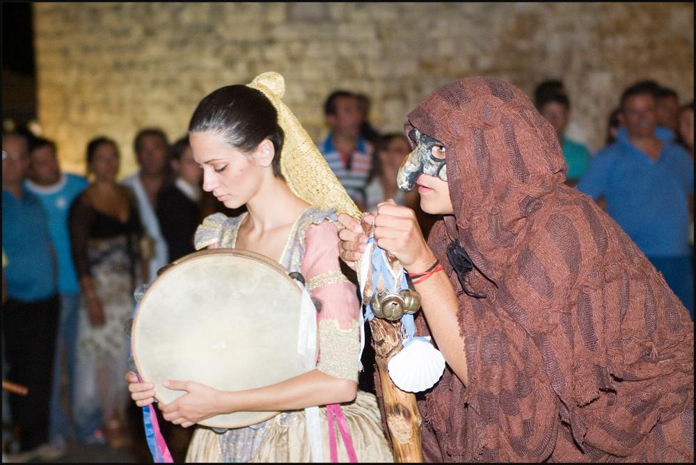 L'immagine digitale è parte di una raccolta fotografica che documenta la seconda edizione della Festa di San Rocco in Contea svoltasi nel 2007 a Conversano e curata dall' associazione culturale “I Collegia”. In primo piano il ritratto di due giovani figuranti in costume di epoca medievale impostati su uno sfondo sfocato in cui si riconosce il pubblico spettatore della parata storica. Il dolce volto della suonatrice di tamburello contrasta sensibilmente con il misterioso personaggio al suo fianco. Il viso nascosto dietro una maschera scura, il corpo leggermente contratto in avanti e avvolto da un mantello in lino marrone, il bastone su cui si poggia e i tre campanellini ad esso attaccati, fanno pensare alla tradizionale rappresentazione del medico della peste, epidemia c...