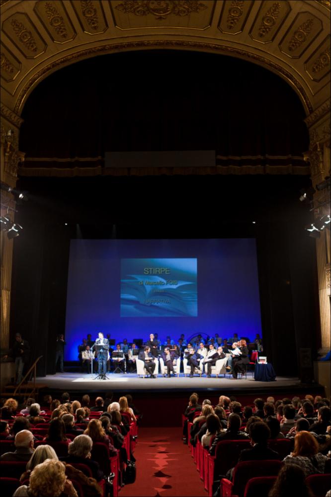 L'immagine digitale cattura uno dei momenti principali della serata di premiazione del Premio Letterario Città di Bari, svoltasi il 21 dicembre 2010, alle ore 19:30, presso il teatro Piccinni di Bari, durante la quale furono presentati tutti i libri in gara. L'edizione 2010 del Premio, alla sua XIII edizione, ebbe il pregio di esaltare le opere letterarie degli scrittori in finale, coinvolgendo attivamente la cittadinanza e spronandola alla lettura e all'ascolto dei brani. Un elemento di novità che, insieme alla giuria di esperti ed una popolare, alle azioni teatrali di Fibre Parallele con "statue parlanti" che hanno letto i libri in molte vie del centro cittadino, ha caratterizzato la XIII edizione del Premio Letterario, dedicato a Pinuccio Tatarella, per questa edizio...
