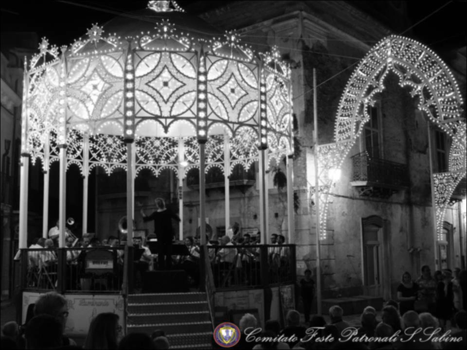 Questo scatto fotografico, gentilmente offerto dal Comitato Festa Patronale di San Sabino, cattura un momento particolarmente suggestivo del concerto bandistico organizzato in occasione della Festa a Torremaggiore nel giugno 2018.
Tra i musicisti e le luminarie che illuminano la piazza con giochi di luce e colori, si diffonde un’atmosfera vibrante di festa e devozione popolare.
La festa patronale di San Sabino, che si celebra ogni anno a Torremaggiore (provincia di Foggia) nel mese di giugno, rappresenta una delle più antiche e radicate tradizioni locali. San Sabino, vescovo e patrono della comunità, fu proclamato tale nel 1216 attraverso una bolla papale di Onorio III. Sotto la sua protezione, generazioni di torremaggioresi si sono succedute, tramandando non solo un pr...