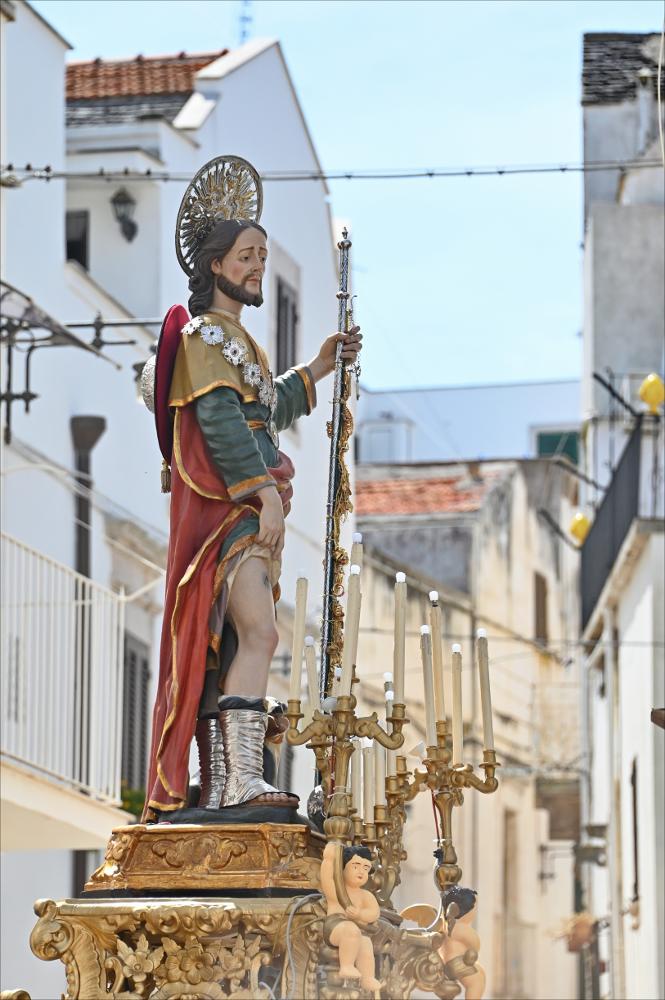 La statua lignea di San Rocco, venerata a Noci, esce in processione. Rappresenta il Santo pellegrino con il bastone, il mantello, lo zaino da pellegrino o bisaccia con la copertura a falda, e il cagnolino ai piedi: elementi che sottolineano la sua vita di carità, il pellegrinaggio e la protezione verso i malati.<div><br>
La festa patronale si celebra tradizionalmente il 16 agosto, giorno dedicato al Santo, mentre la solennità liturgica propria della comunità nocese ricorre nella prima domenica di settembre, cuore delle celebrazioni religiose e civili.
La devozione a San Rocco a Noci nasce agli inizi del Cinquecento, in un contesto segnato dal timore della peste che colpiva i territori vicini. Pur non essendo storicamente devastata dal contagio, la comunità sviluppò una ...
