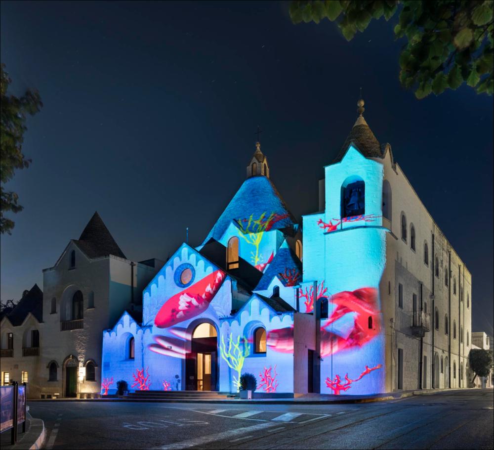 Lo scatto fotografico riprende la proiezione interattiva “Aquarium”, dello studio portoghese Ocubo, che trasforma gli edifici in acquari giganti. La Chiesa di Sant’Antonio di Alberobello ha ospitato il gigantesco acquario interattivo dove i visitatori hanno potuto realizzare pesci personalizzati attraverso un apposito pannello touch screen.
La proiezione è stata realizzata nell’ambito della seconda edizione del Life - Alberobello Light Festival 2017 che ha illuminato il centro cittadino e alcuni dei suoi luoghi più caratteristici dal 30 settembre all’8 ottobre.
Il Festival è organizzato da Lightcones e Agit - Associazione Giovani Imprenditori del Turismo, responsabili rispettivamente della direzione artistica e della direzione organizzativa del Festival. L’edizione 2017...