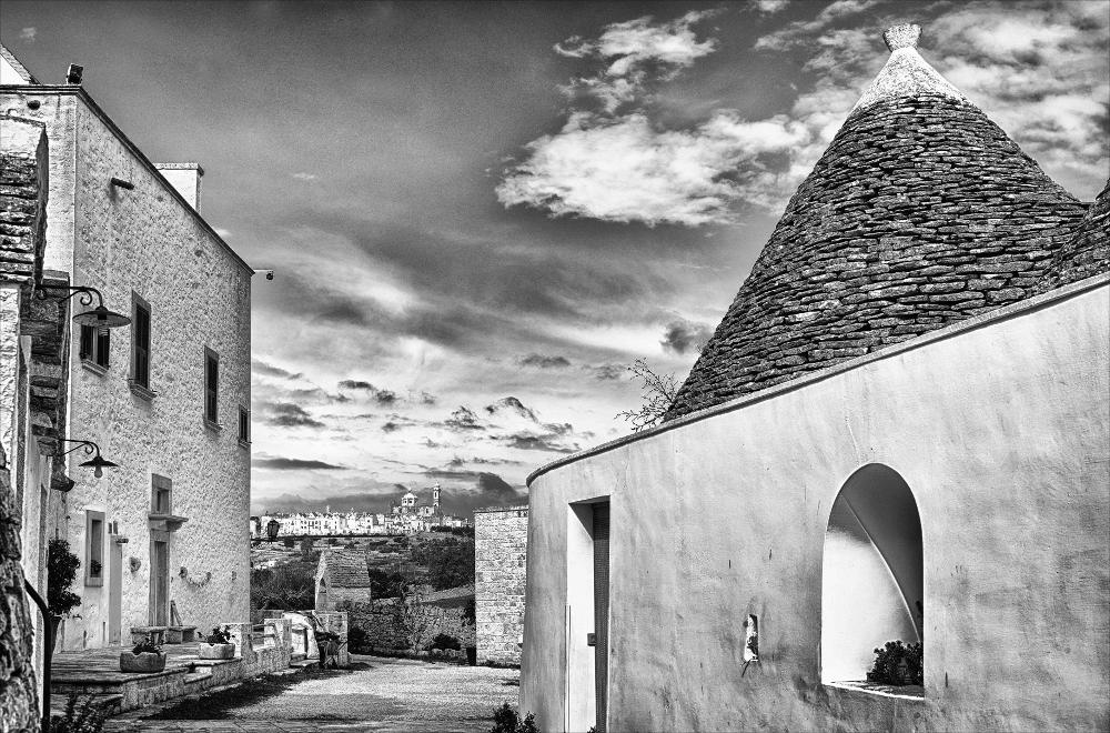 L'istantanea in oggetto, fa parte di una più ampia raccolta di fotografie realizzate per il progetto di censimento di 248 masserie pugliesi avviato da Giancarlo Chielli, professore del corso in Beni Culturali e Ambientali presso l'Accademia di Belle Arti di Bari. Il lavoro svolto ha dato vita al volume “Le masserie di Puglia come Bene Culturale” in cui sono presenti alcuni scatti di masserie pugliesi tra cui questa presa in esame. La fotografia recupera uno scorcio della masseria Aprile utilizzando una inquadratura grandangolare che consente all'immagine di estendersi in profondità e di individuare alcuni ambienti del complesso rurale destinati in origini a diverse funzione. In fondo alla foto si intravede in lontananza il paesaggio del centro storico di Locorotondo, st...