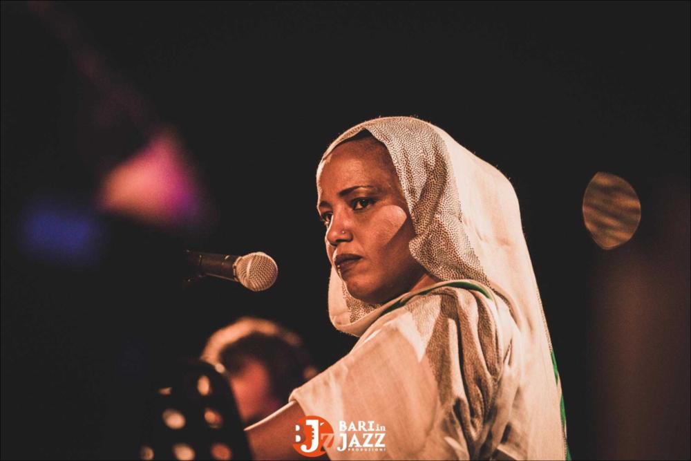 "Bari in Jazz" è un Festival Metropolitano con cadenza annuale, organizzato da Abusuan in Puglia, presenta concerti con ospiti nazionali ed internazionali. L’edizione 2018 del Festival Metropolitano "Bari in Jazz", la quattordicesima, è stata interamente dedicata alle figure femminili nella musica e nel jazz, nel canto, nella composizione e nell’arrangiamento, nell’uso dei vari strumenti e stili musicali. Come ogni anno il Festival Metropolitano Bari in Jazz, luogo d’incontro della musica jazz con altri generi musicali, offre una vetrina d’eccellenza alle grandi artiste e a quelle emergenti, promuovendo anche nuove creazioni. La fotografia in oggetto pone in risalto, al centro, la cantante; vestita in abiti tipici si distacca cromaticamente dal fondo scuro rendendo il r...