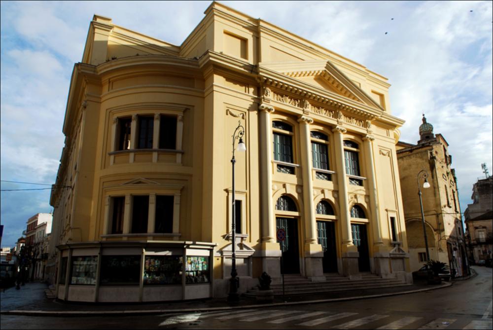 L'immagine è parte della campagna fotografica realizzata dalla fotografa Margherita Mirabella per Teatro Pubblico Pugliese che, con i suoi scatti, ha ripreso le eleganti architetture e i rifiniti dettagli di alcuni dei più bei teatri pugliesi e di alcuni spettacoli teatrali portati in scena in questi ultimi anni. L'immagine ci restituisce l'imponente e rigorosa facciata neoclassica del Teatro Comunale "Giuseppe Verdi" di San Severo, riconosciuto come la più grande struttura teatrale di Capitanata e terzo teatro all'italiana più importante di Puglia dopo il Petruzzelli di Bari e il Politeama Greco di Lecce. Il monumento, originariamente denominato teatro Comunale del Littorio, viene progettato dall'architetto Cesare Bazzani rispettando a pieno i canoni stilistici dell'ar...