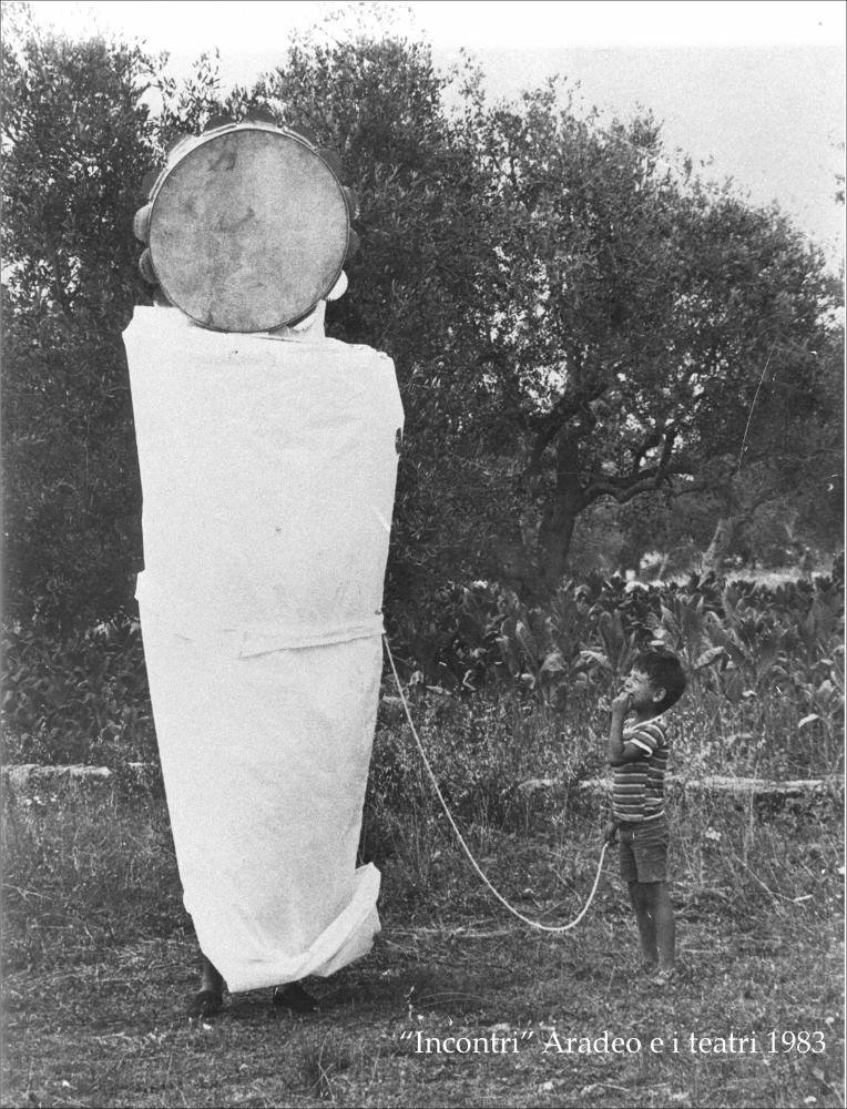 L'immagine digitale immortala un piccolo momento della rassegna “Aradeo e i Teatri” inaugurata nello stesso anno, 1983, con il titolo “Incontri”, un appuntamento con il teatro sperimentale. La manifestazione fu organizzata dal circolo culturale “S.M. Ejzenstein” per colmare, attraverso la messa in scena di spettacoli di teatro e danza, la mancanza di confronto fra culture differenti molto sentita dalla cittadinanza di Aradeo. Nello specifico si osserva un “tamburello gigante”, strumento principe della pizzica tipico della tradizione salentina. La fotografia mette in luce come da oggetto inanimato il tamburello viene innalzato a maschera, volto di un figurante completamente ricoperto da un costume bianco dal quale restano visibili solo i piedi. In basso è presente un pic...