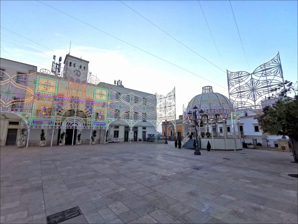 Lo scatto ritrae la piazza Moro della città di Locorotondo mentre sono in corso gli allestimenti delle Luminarie in occasione dei festeggiamenti in onore del Compatrono e Protettore San Rocco nel mese di agosto 2024.
La Festa Patronale di San Rocco si svolge dal 6 al 17 agosto, con riti religiosi e iniziative civili. Il momento centrale è tra il 15 e il 17 agosto, con quattro processioni principali: la traslazione della statua (15), due processioni il 16 (mattutina storica e serale di gala) e la suggestiva processione serale “aux flambeaux” del 17, che conclude la festa.
Sul piano civile, l’apertura simbolica è l’alba del 16 agosto con le bande che suonano per le stradine della città la poesia in vernacolo “A’ Diènè”, vera e propria colonna sonora della festa; molto att...