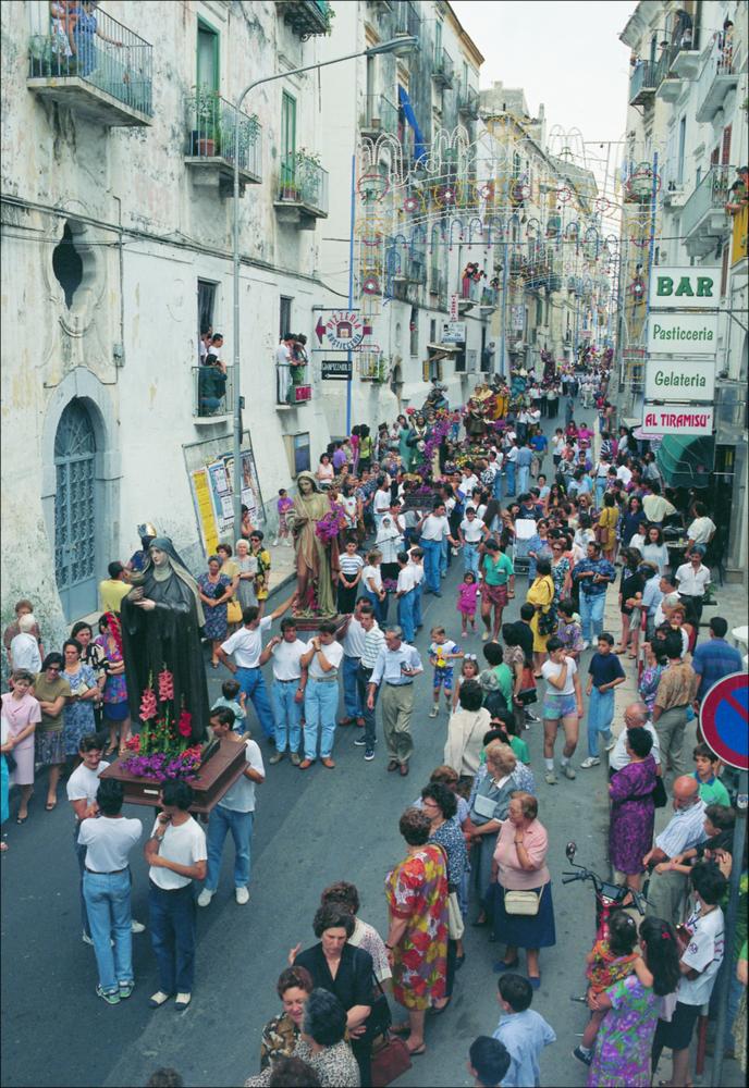 La fotografia è parte della ricca raccolta fotografica “Puglia per tutte le Stagioni” vol. 2, prodotta dall'Assessorato al Turismo e Industria Alberghiera della Regione Puglia e creata per raccontare la Puglia attraverso immagini diverse suddivise in 5 settori principali: Città e Paesi, Settimana Santa, Territorio, Feste – Sagre – Tradizioni, Mercati – Prodotti – Cucina. L'immagine cattura un momento della processione dei Santi in occasione delle festa patronale di Rodi Garganico, che ricorre ogni anno il 2 e il 3 luglio. In questi giorni, dedicati ai festeggiamenti in onore della patrona Maria Santissima della Libera e di San Cristoforo, per le vie della città si alternano processioni e momenti religiosi a mercatini ed eventi culturali. La tradizione vuole che, agli in...