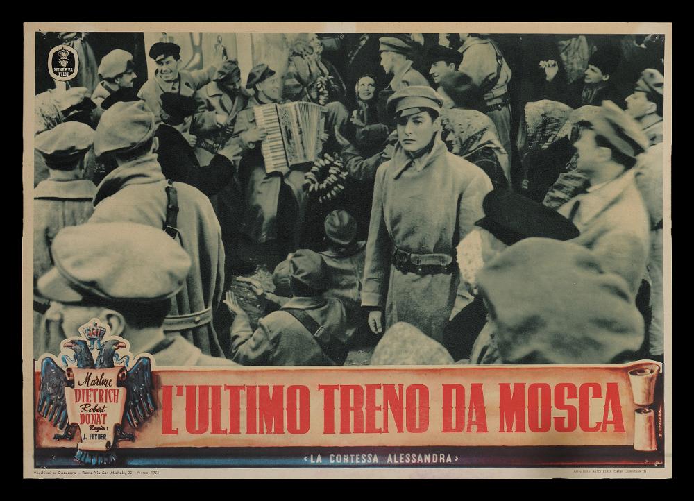 L'immagine riproduce uno dei soggetti delle fotobuste conservate presso la Mediateca Regionale Pugliese (vedi Note). Si tratta, nello specifico di una delle fotobuste del film in bianco e nero “L’ultimo treno da Mosca”, in Italia distribuito anche con il titolo “La contessa Alessandra”, film del 1937 per la regia di Jacques Feyder. La fotografia immortala una scena all’aperto, nel quale sono riconoscibili dei soldati, ma l’attenzione ricade sulla bellissima Marlene Dietrich, la contessa Alessandra Vladinoff, colta in un momento di grande intensità. Al di sotto dell’immagine possiamo notare un cartiglio contenente il titolo a caratteri cubitali e rossi e lo stemma dell’impero russo, l’aquila bicipite, spesso utilizzata in araldica anche come stemma di altre nazioni. La f...