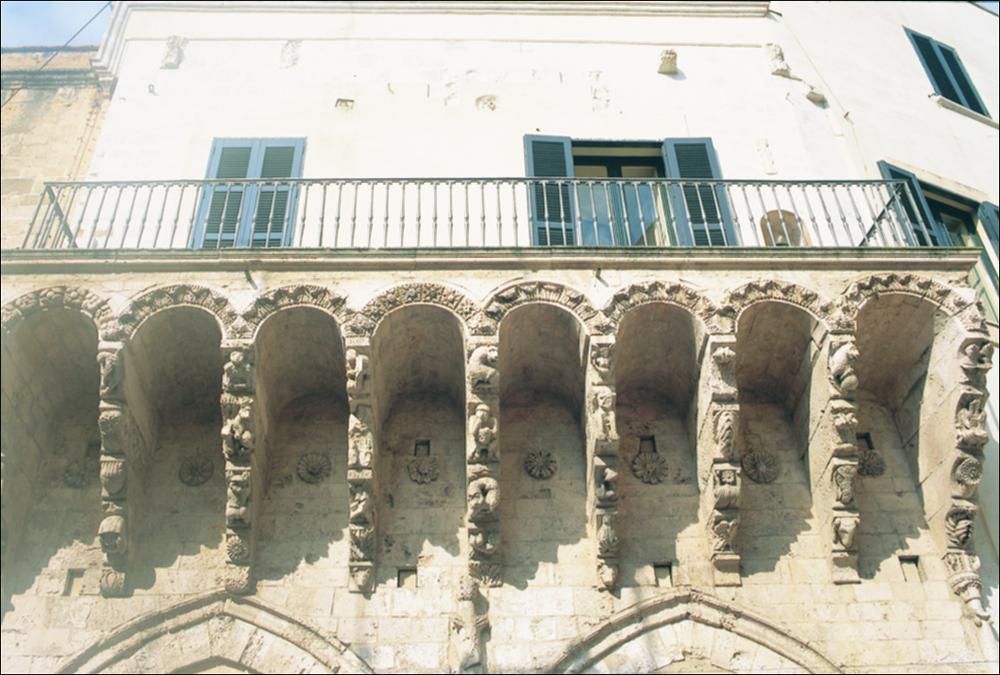 La fotografia è parte della ricca raccolta fotografica “Puglia per tutte le Stagioni” vol. 2, prodotta dall'Assessorato al Turismo e Industria Alberghiera della Regione Puglia, per raccontare la Puglia attraverso immagini suddivise in 5 settori principali: Città e Paesi, Settimana Santa, Territorio, Feste – Sagre – Tradizioni, Mercati – Prodotti – Cucina. Si tratta di una ripresa dal basso della Loggia Balsamo, parte dell'omonimo palazzo storico, situato a Brindisi in Piazza Duomo nei pressi della Cattedrale. Il balcone poggia su una serie di mensole con archi a tutto sesto, decorati da motivi squisitamente naturalistici, come fiori e foglie, tra i quali spiccano alcune figurine. Le mensole sono finemente decorate da sculturine allegoriche, figure bizzarre e animali fan...