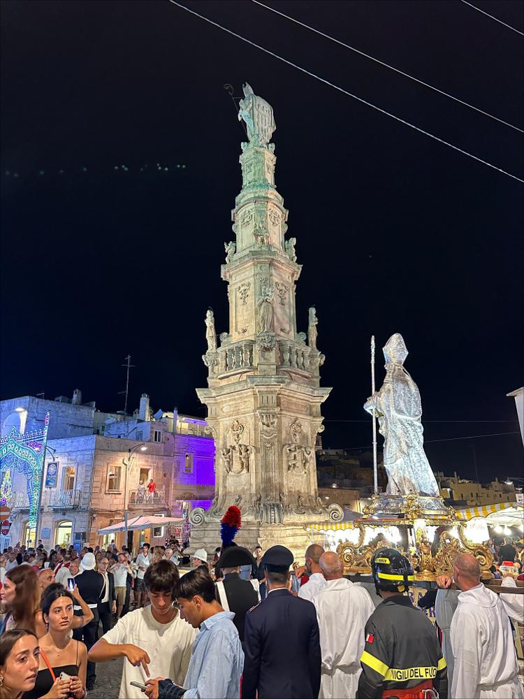 <div class="ql-editor"><div>L’immagine ritrae una suggestiva scena notturna nel centro storico di Ostuni i festeggiamenti in onore di Sant'Oronzo. In primo piano spicca la <strong>Colonna di Sant’Oronzo</strong>, monumento barocco che domina Piazza della Libertà, mentre accanto il simulacro argenteo del Santo viene portato in processione tra i fedeli, le autorità e i membri delle forze dell’ordine.</div><div>Le luminarie e la folla evidenziano l’atmosfera festiva e partecipativa tipica delle celebrazioni patronali pugliesi, dove devozione, storia e vita sociale si intrecciano.</div><div><br></div><div>A Ostuni, ogni anno dal 25 al 27 agosto, si rinnova uno degli appuntamenti più sentiti dalla comunità: la Cavalcata in onore di...