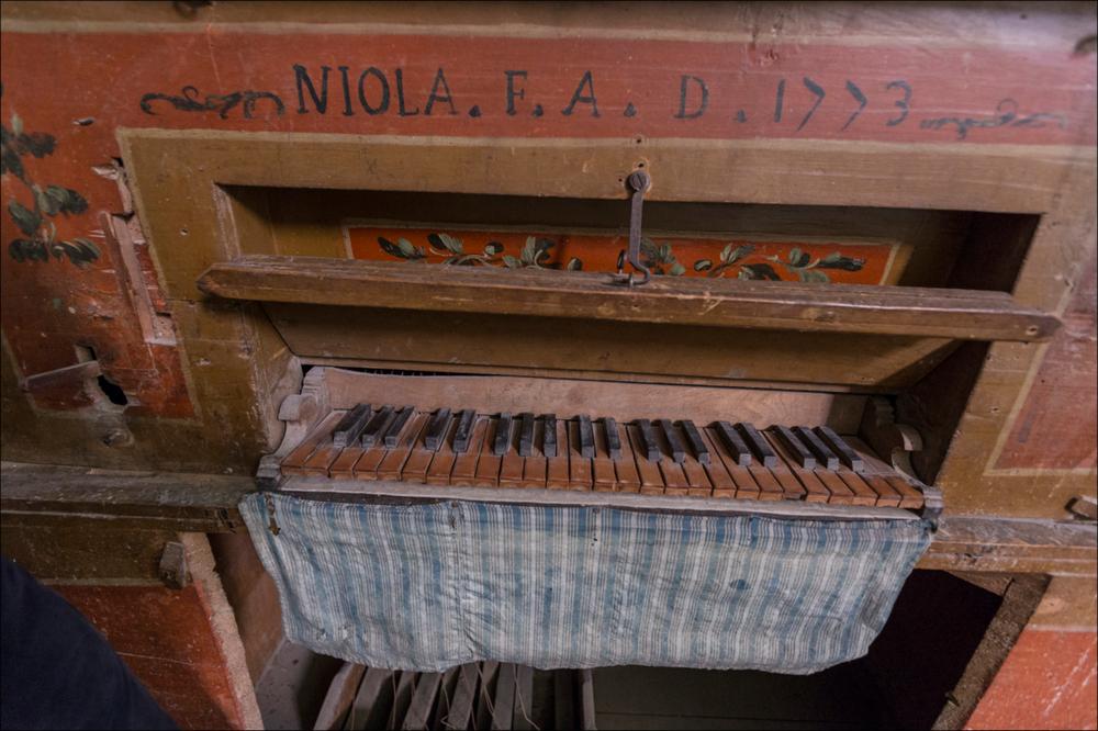 L'immagine cattura in primo piano un dettaglio dell’organo, collocato nella chiesa di S. Leonardo a Manduria. La chiesa di piccole dimensione fu costruita su una preesistente cappella intitolata alla Madonna di Costantinopoli. Dedicata a S. Leonardo dal 1621, venne ricostruita a partire dal 1702, grazie ai contribuiti dei confratelli. La facciata molto semplice presenta doppie paraste ai lati e un fastigio a volute, collocato su una trabeazione continua, sormontato da una croce. Il portale centrale, unico ingresso, vede al centro all’interno una nicchia con la statua di San Leonardo, ai lati San Cristoforo e San Raffaele. L’interno ad aula unica presenta sull’altare maggiore, realizzato in pietra leccese e con colonne tortili, la tela della Madonna di Costantinopoli rea...
