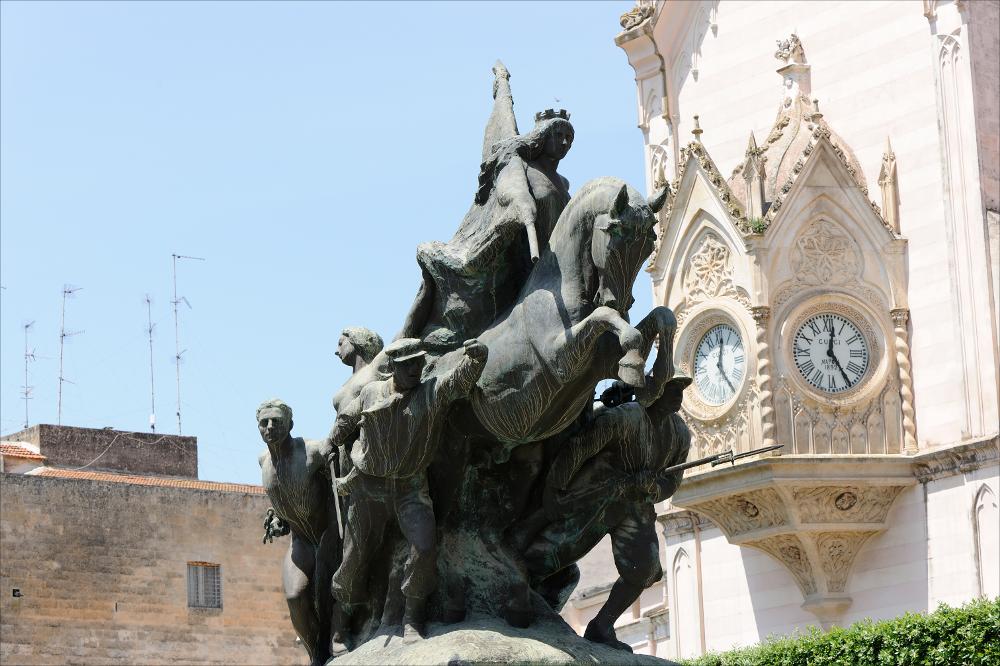 L'immagine digitale mostra il Monumento ai Caduti a Gravina in Puglia, opera dello scultore Angelo Galli, sito nella Villa Comunale. Protagonista assoluta della maestosa opera bronzea, è la donna, personificazione dell'Italia, in sella ad un cavallo immortalato nell'atto di impennarsi. In basso, ai lati del cavallo sono presenti dei soldati armati, scolpiti nel momento che precede la carica durante il combattimento, si può notare infatti come un milite sia ripreso mentre suona la tromba per incitare il gruppo alla battaglia, caricando di grande dinamismo la scena. Nel secondo gruppo di soldati si può notare la presenza di un prigioniero, metafora del popolo che si ribella all'oppressore, riconoscibile dalle catene tagliate ancora legate ai polsi. A fare da sfondo alla s...