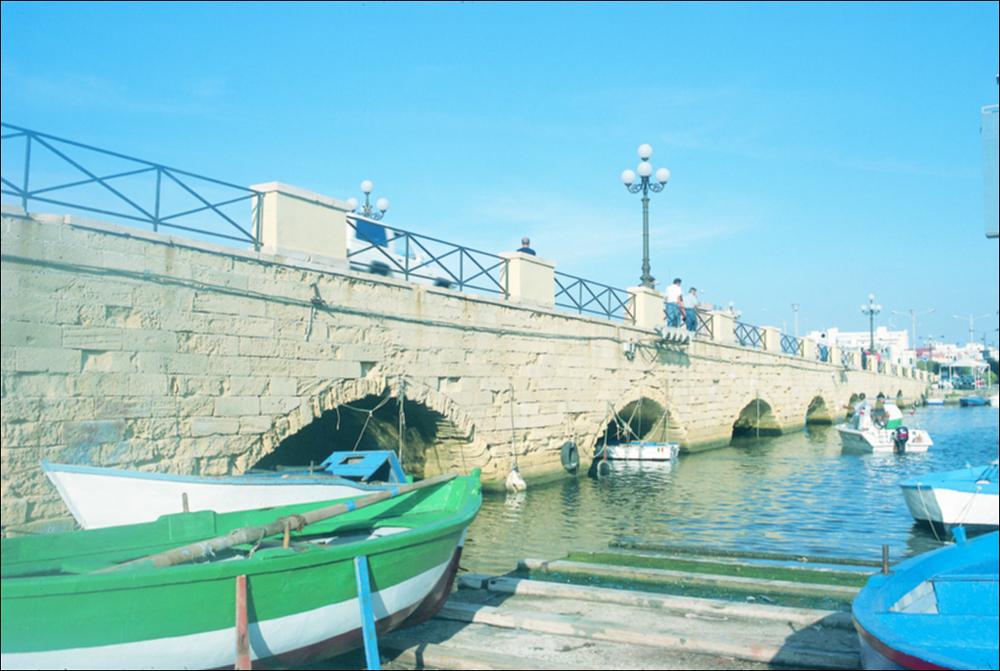 La fotografia è parte della ricca raccolta fotografica “Puglia per tutte le Stagioni” vol. 2, prodotta dall'Assessorato al Turismo e Industria Alberghiera della Regione Puglia, per raccontare la Puglia attraverso immagini suddivise in 5 settori principali: Città e Paesi, Settimana Santa, Territorio, Feste – Sagre – Tradizioni, Mercati – Prodotti – Cucina. L'immagine immortala uno scorcio del Ponte civico di Gallipoli che collega il centro storico, caratterizzato da vie strette e tortuose, chiese, antichi edifici storici, civili e militari, alla parte nuova della città. La storia del ponte di Gallipoli risale al 1484, quando la Città passò in mano ai Veneziani che. per difendere l'isola dagli attacchi nemici decisero di tagliare l'istmo che univa il borgo antico alla par...