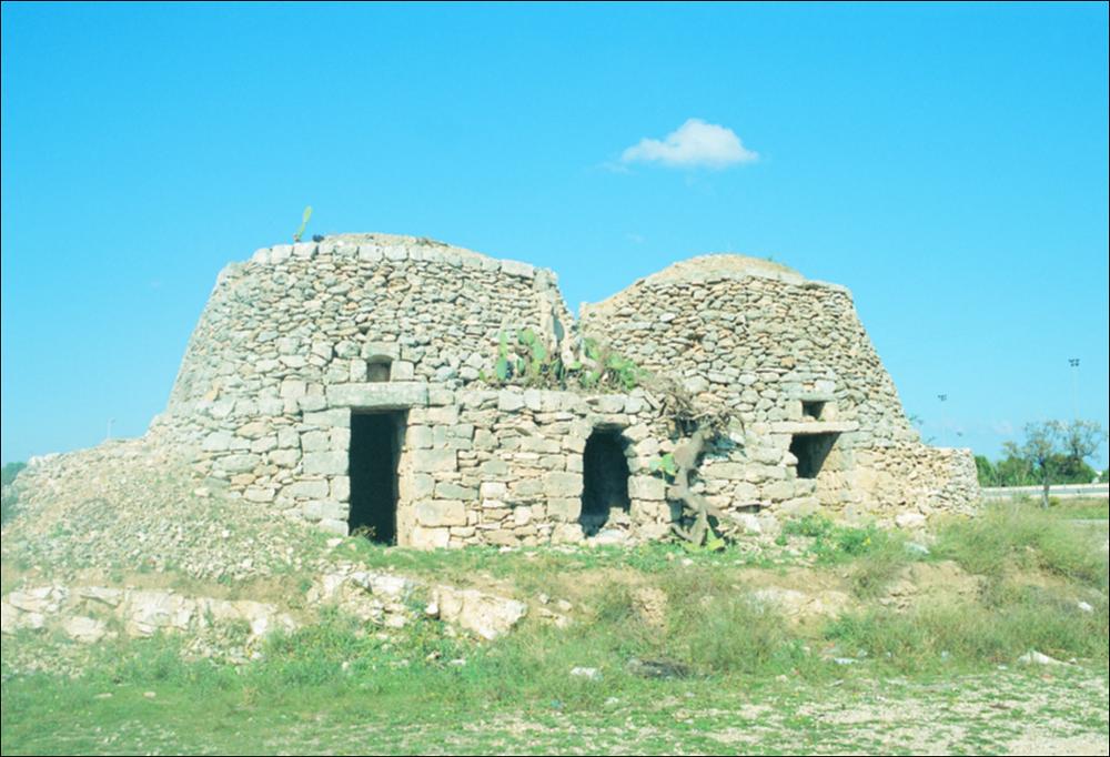 L'immagine è parte della ricca raccolta fotografica “Puglia per tutte le Stagioni” vol. 2, prodotta dall'Assessorato al Turismo e Industria Alberghiera della Regione Puglia per raccontare la Puglia attraverso immagini suddivise in 5 settori principali: Città e Paesi, Settimana Santa, Territorio, Feste – Sagre – Tradizioni, Mercati – Prodotti – Cucina. La fotografia mostra due trulli affiancati, deteriorati dagli agenti atmosferici, strutture architettoniche caratteristiche del paesaggio pugliese fin dal I millennio a.C. I trulli sono delle costruzioni a base circolare, in pietra a secco, della zona centro meridionale della Puglia, note per la loro cupola conica, tipicamente realizzate dai contadini e utilizzate come abitazioni permanenti o ricoveri temporanei. La loro a...
