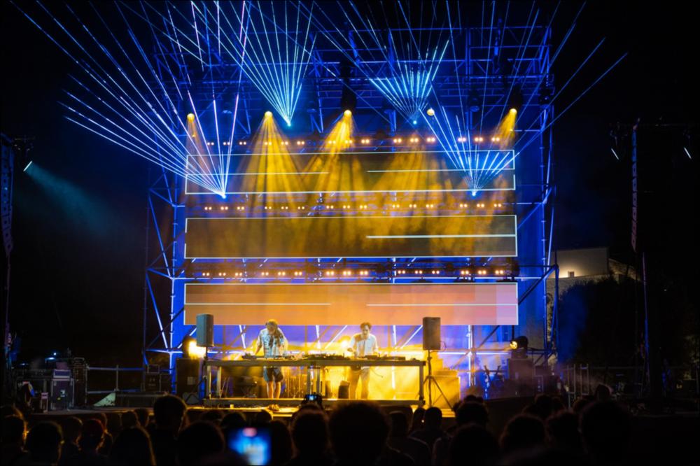 Nello scatto fotografico giochi di illuminotecnica sul palcoscenico che accoglie l’esibizione dei Nu Genea durante la serata di VIVA! Festival 2021 del 6 agosto. I Nu Genea, Massimo Di Lena e Lucio Aquilina, sono due musicisti/dj napoletani di stanza a Berlino. Nel loro percorso artistico e musicale partono dalle loro radici napoletane per esplorare e sperimentare nuovi e più lontani mondi musicali fino a elaborare un’idea di musica groovy impregnata di disco, funk, elettronica, dub, folk e molto altro.
Lo show "Neapolitan Middle-Eastern Special" del 6 agosto è stato uno evento speciale appositamente prodotto per VIVA! e dedicato ai suoni del Mediterraneo e del Medio Oriente....