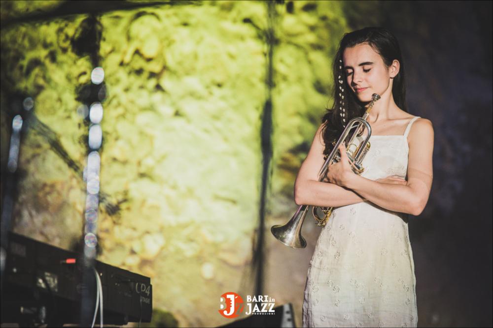 "Bari in Jazz" è un Festival Metropolitano con cadenza annuale, organizzato da Abusuan in Puglia, presenta concerti con ospiti nazionali ed internazionali. L’edizione 2018 del Festival Metropolitano "Bari in Jazz", la quattordicesima, è stata interamente dedicata alle figure femminili nella musica e nel jazz, nel canto, nella composizione e nell’arrangiamento, nell’uso dei vari strumenti e stili musicali. Come ogni anno il Festival Metropolitano Bari in Jazz, luogo d’incontro della musica jazz con altri generi musicali, offre una vetrina d’eccellenza alle grandi artiste e a quelle emergenti, promuovendo anche nuove creazioni. La fotografia in oggetto mostra un momento delle Esibizioni musicali "Under 35", in cui una ragazza, in età adolescenziale, stringe una tromba, in...