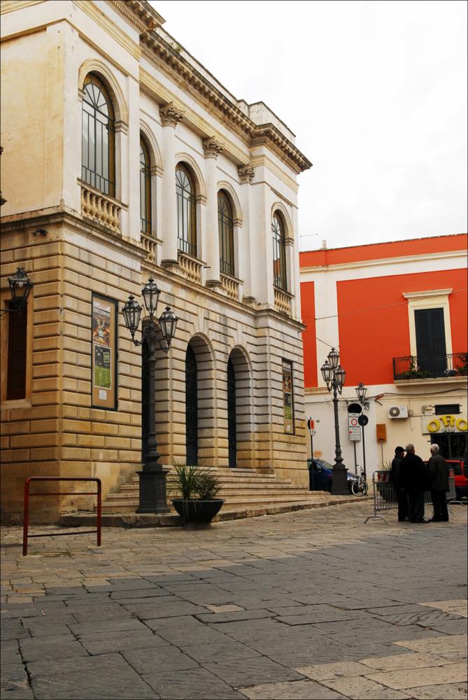 L'immagine è parte della campagna fotografica realizzata dalla fotografa Margherita Mirabella per Teatro Pubblico Pugliese, che ha ripreso nei suoi scatti le eleganti architetture e i rifiniti dettagli di alcuni dei più bei teatri pugliesi e alcuni degli spettacoli teatrali portati in scena in questi ultimi anni. Da una prospettiva laterale, l'autrice cattura la facciata del Teatro Comunale di Nardò, situato in Via Mauro Manieri, piccolo gioiello culturale ottocentesco della città salentina. Dopo l'Unità d'Italia, l'esigenza di avere un Teatro Comunale per la popolazione locale spinse la Prefettura ad affidare l'incarico per la sua realizzazione. Nel 1893, fu accolto il progetto dell'Ing. Quintino Tarantino e i lavori iniziarono nello stesso anno su alcuni immobili demo...