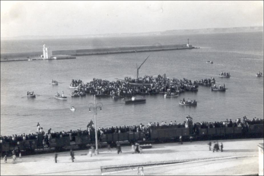 Fotografia storica del 1938 raffigurante "la cuccagna a mare" nel porto di Gallipoli, in occasione dei festeggiamenti in onore di Santa Cristina. Il palo della cuccagna è collocato in mare, al centro del porto, ed è circondato da numerose imbarcazioni. Lungo la banchina si osserva una folla compatta di spettatori che assiste all’evento da lontano.

Lo scatto documenta l’utilizzo del bacino portuale come spazio della celebrazione. La fotografia testimonia la continuità storica della cuccagna a mare nella prima metà del XX secolo.
La festa patronale di Santa Cristina, protettrice della città di Gallipoli, si svolge annualmente nei giorni 23, 24 e 25 luglio e rappresenta uno dei momenti più sentiti della vita religiosa, sociale e c...